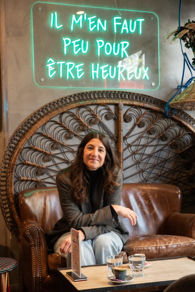 Solene Gentili, interprète en langue des signes, assise dans un bar sour un texte en neon qui proclame : Il m'en faut peu pour être heureux